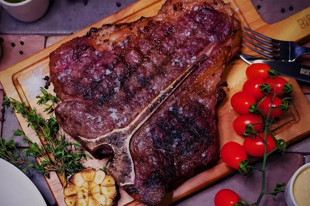 Juicy T‑bone steak with coarse salt, roasted garlic, cherry tomatoes, and fresh herbs served on wooden board at Bistecca Tuscan Steakhouse.