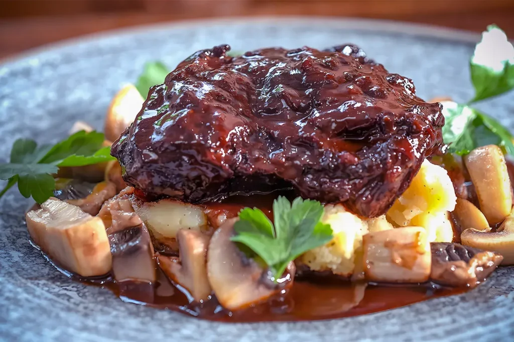 Close-up of a richly glazed beef dish on mashed potatoes, surrounded by sautéed mushrooms and garnished with fresh parsley on a grey plate.