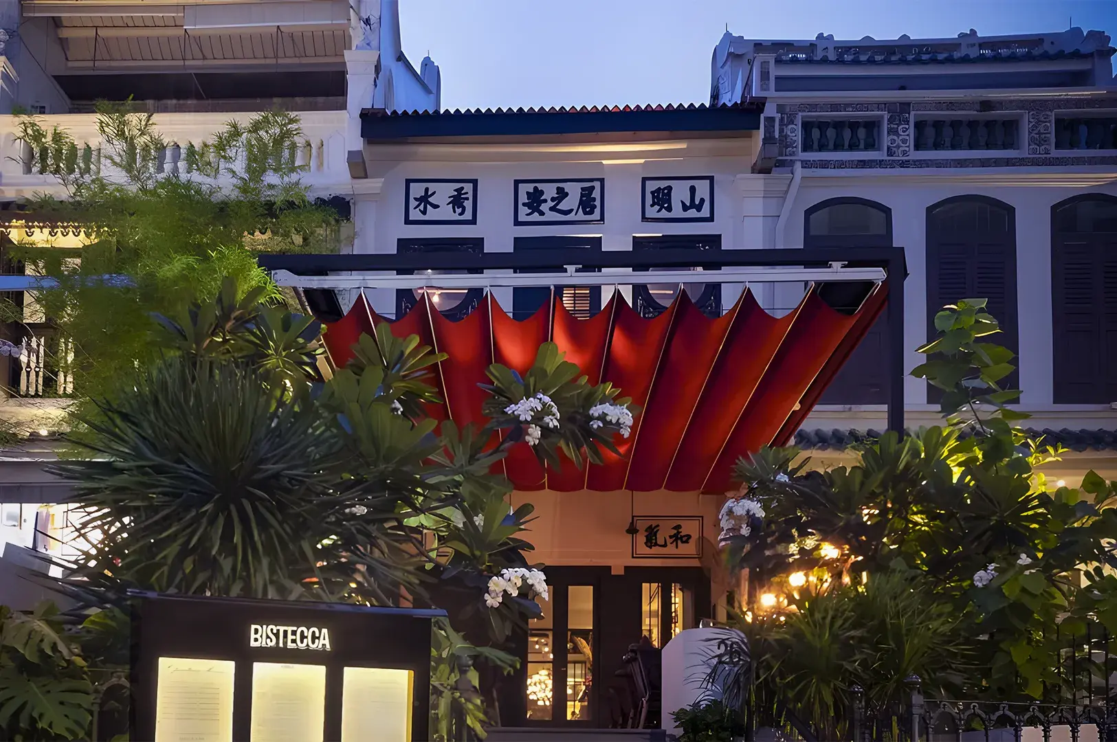 Elegant exterior of Bistecca Tuscan Steakhouse featuring a red canopy, lush greenery, and traditional shophouse architecture in Singapore.
