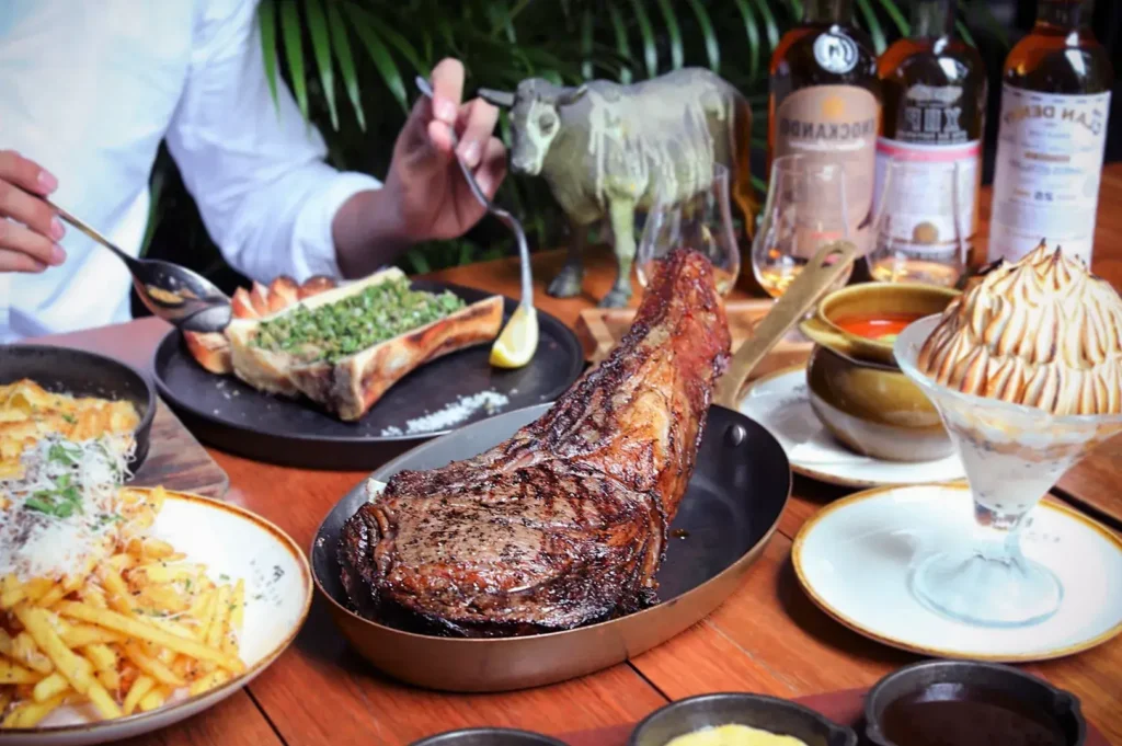 A lavish spread at Bedrock Bar & Grill featuring a large Tomahawk steak, roasted bone marrow, truffle fries, and a flaming Baked Alaska dessert.