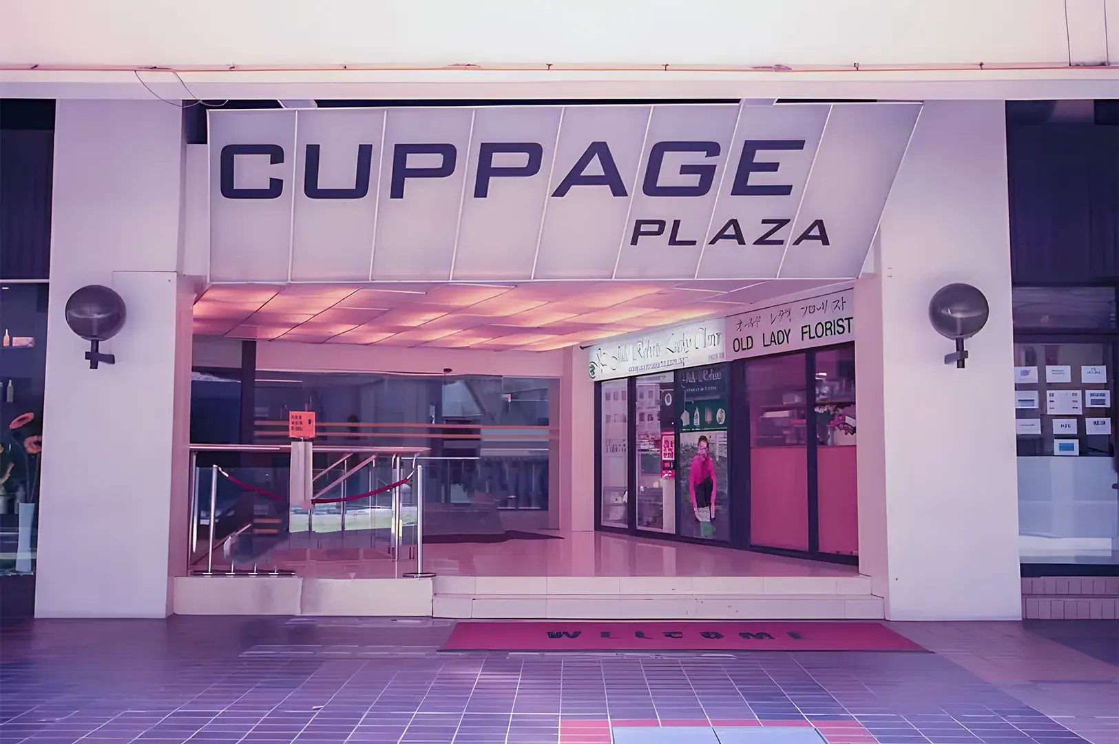 Entrance to Cuppage Plaza, featuring a large sign above a glass doorway. A red welcome mat is on the tiled floor, and shops are visible inside.