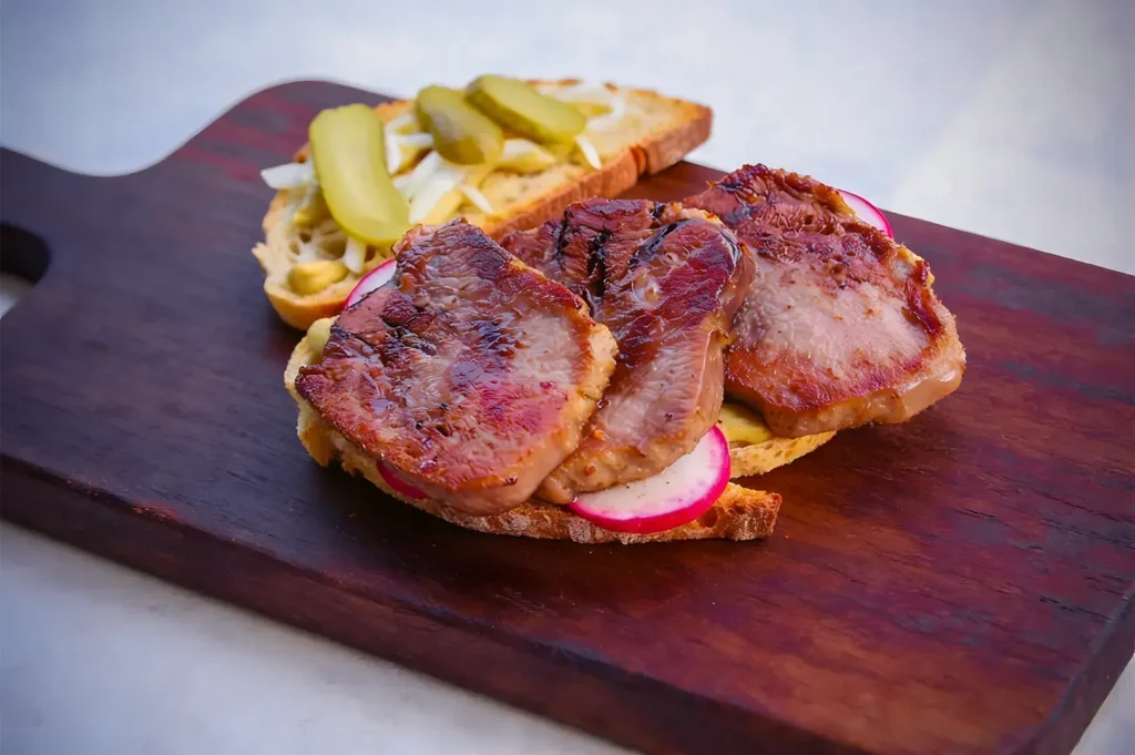 Open sandwich on a wooden board with grilled meat, pickles, and radish slices on sliced bread, creating a rustic, hearty presentation.