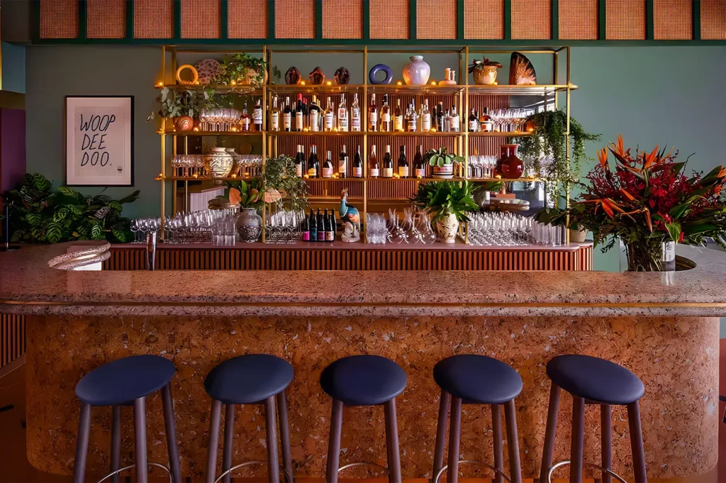 Stylish bar interior with cork counter, five blue stools, and colorful glassware. Green wall with art, lush plants, and warm lighting creates a cozy atmosphere.