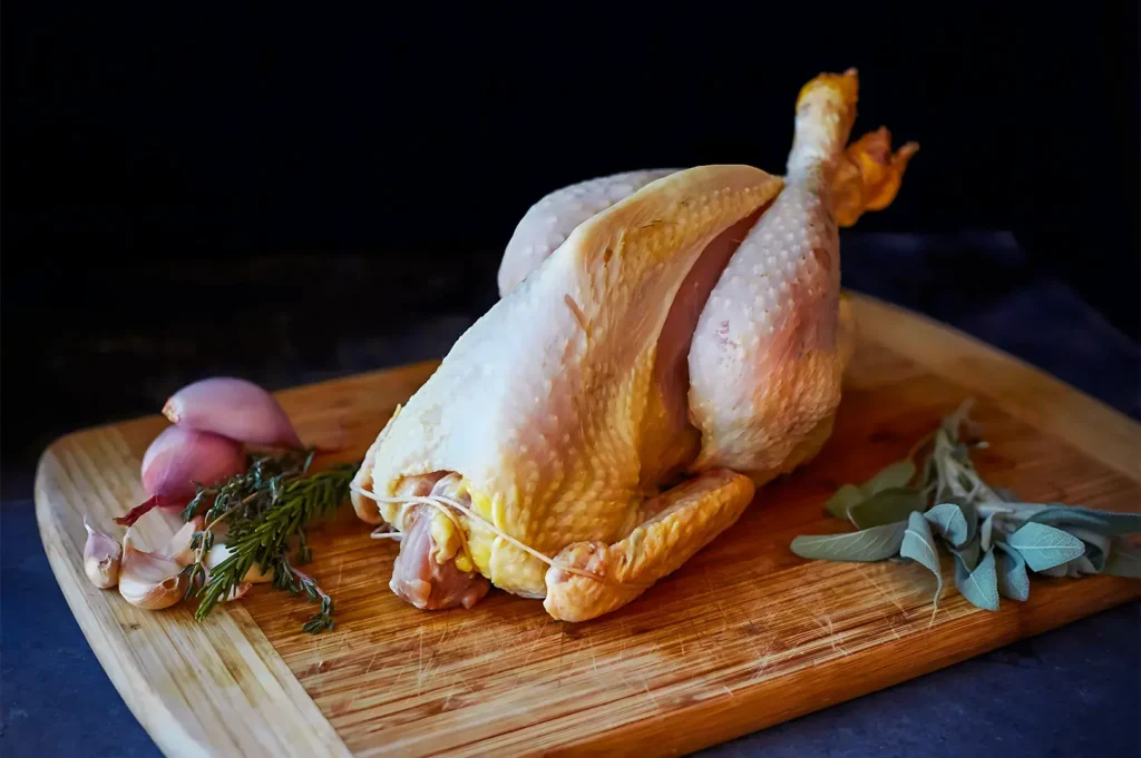 Raw whole chicken trussed on a wooden board with fresh garlic, shallots, rosemary, and sage. The setting suggests preparation for cooking.