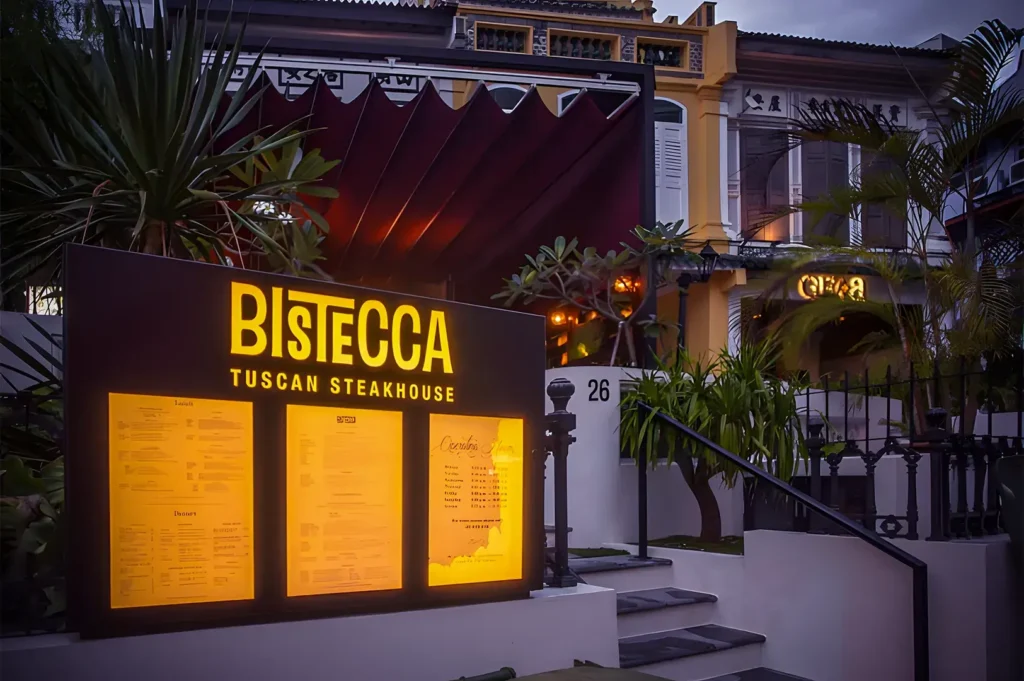 Illuminated Bistecca Tuscan Steakhouse sign with glowing yellow menu display and classic shophouse façade surrounded by tropical plants.