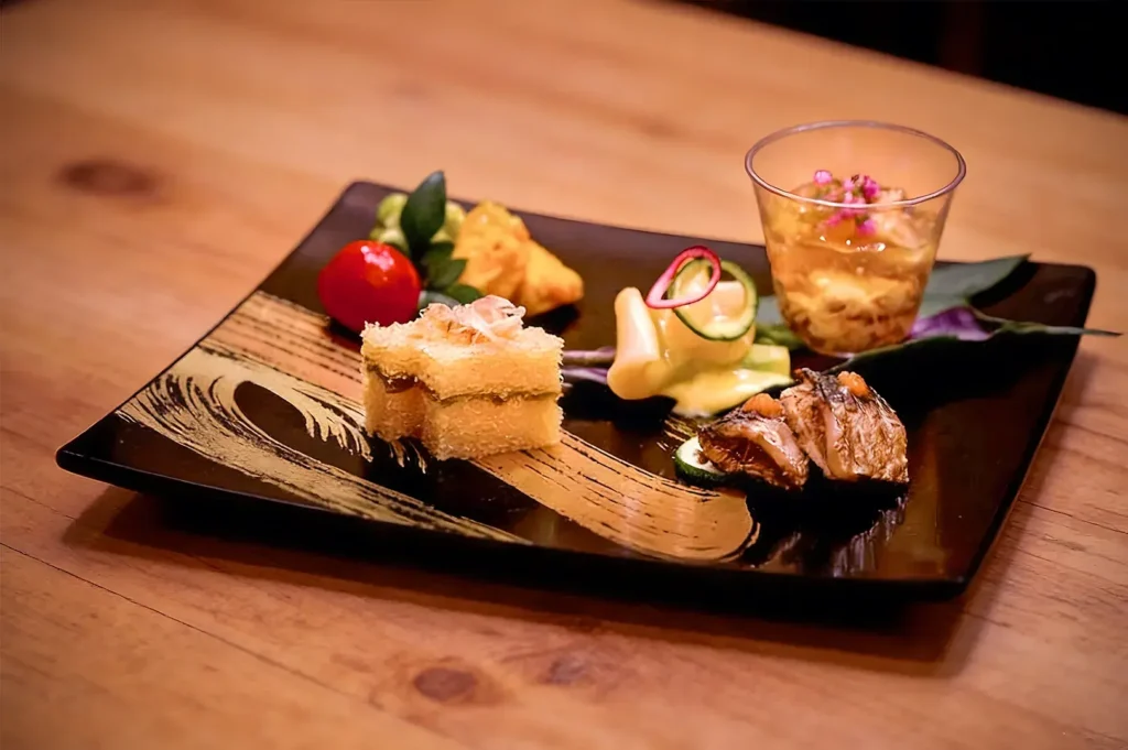 A beautifully arranged gourmet dish on a black plate with artistic patterns. Includes layered cake, grilled meat, a red fruit, and a garnish of edible flowers.