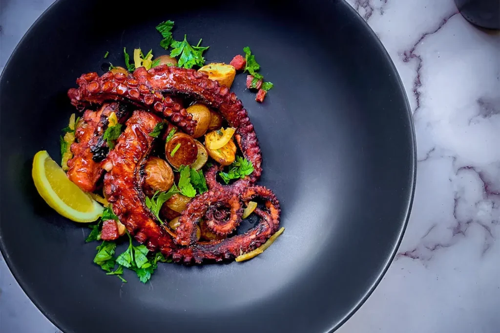 Grilled octopus tentacles on a black plate with roasted potatoes, fresh parsley, diced bacon, and lemon wedge, on a marble surface.