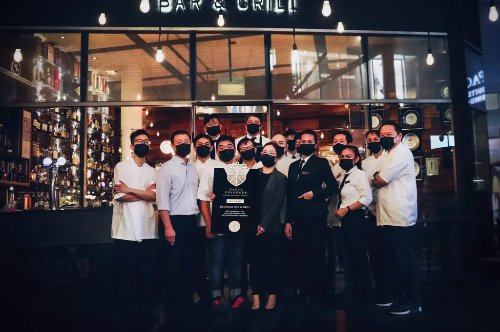The professional culinary and service team of Bedrock Bar & Grill posing with a Haute Grandeur Global Excellence award in front of the bar.