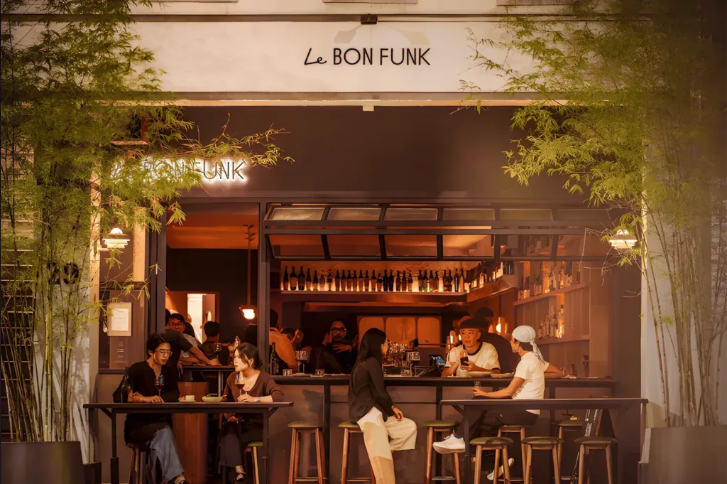 Cozy outdoor seating at Le Bon Funk wine bar with patrons enjoying drinks and conversation. Warm lighting and greenery create an inviting atmosphere.