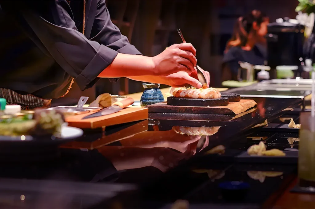 Chef's hands artfully arranging sushi on a black counter, with focus and precision. Warm lighting and reflections create an elegant, inviting atmosphere.