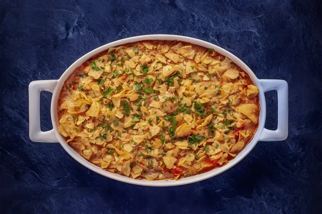 A white oval casserole dish filled with a cheesy chicken tortilla casserole, topped with crispy tortilla chips and garnished with fresh herbs on a dark blue background.