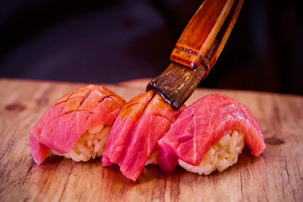 Three pieces of sushi topped with vivid pink tuna are on a wooden board. A brush is glazing them with sauce, highlighting the fresh texture.