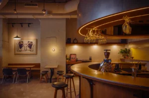 Cozy cafe interior with curved wooden bar, hanging glasses, and warm lighting. Minimalist art on walls and modern seating create a relaxed ambiance.