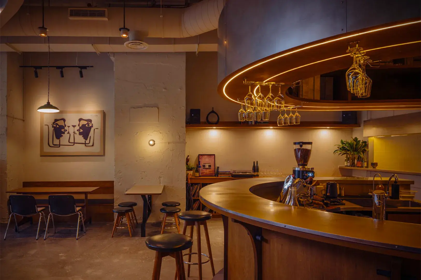 Cozy cafe interior with curved wooden bar, hanging glasses, and warm lighting. Minimalist art on walls and modern seating create a relaxed ambiance.