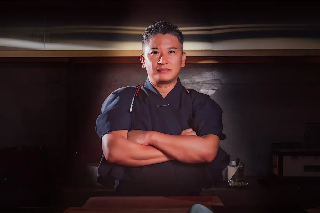 A chef stands confidently with arms crossed in a dimly lit kitchen, wearing a dark uniform. The atmosphere is professional and focused.
