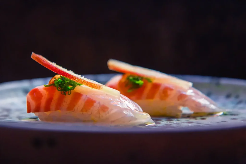 Two pieces of nigiri sushi with salmon slices, apple slivers, and green garnish on rice atop a ceramic plate, lit dramatically against a dark background.