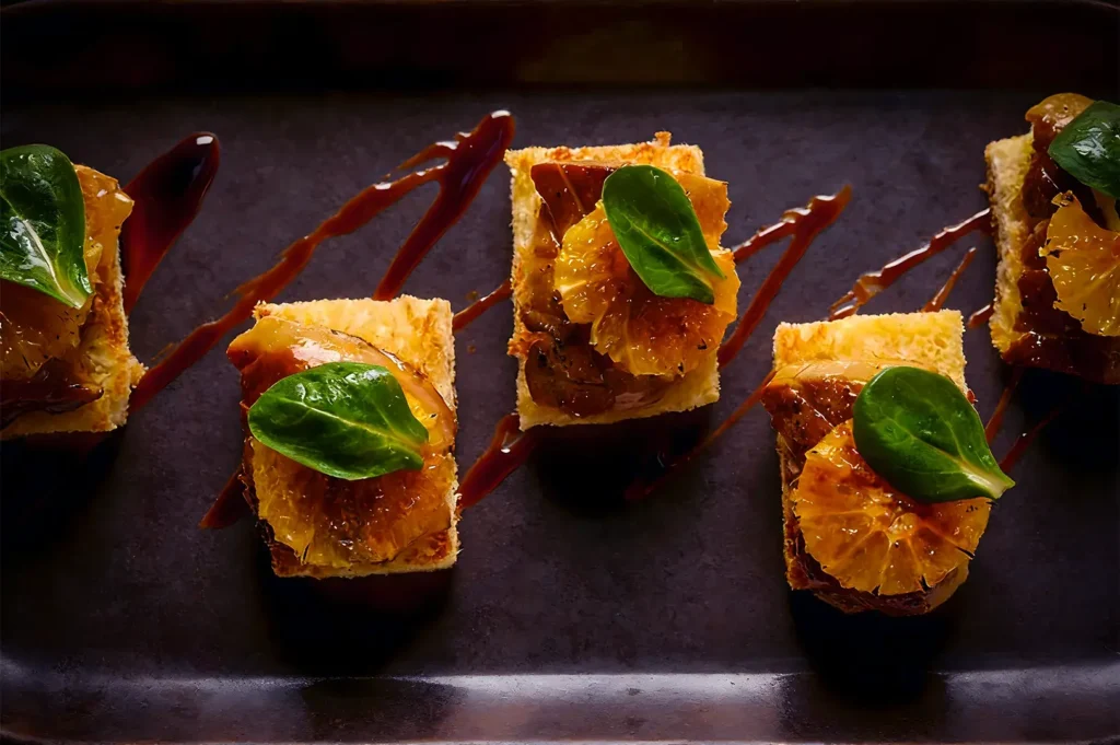 Gourmet appetizers on a dark plate, featuring layered bread, caramelized orange slices, and vibrant green leaves drizzled with a glossy brown sauce.