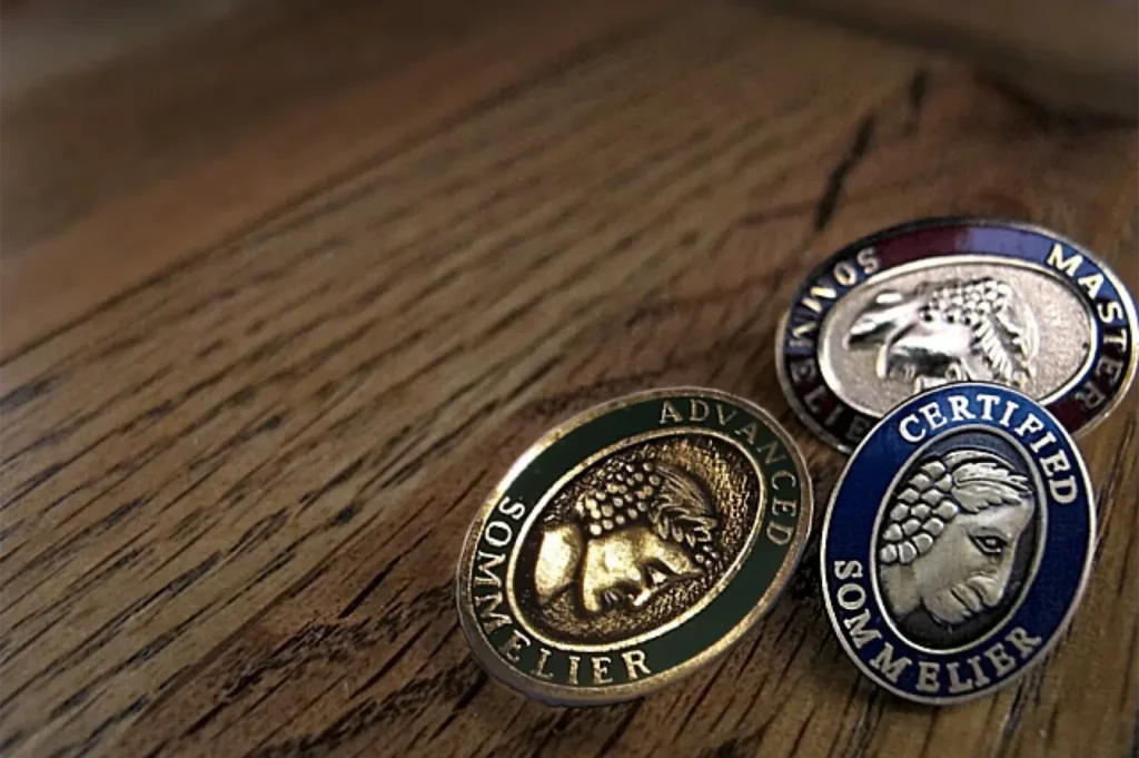 Close-up of three official Court of Master Sommeliers lapel pins—Certified, Advanced, and Master—resting on a wooden surface.