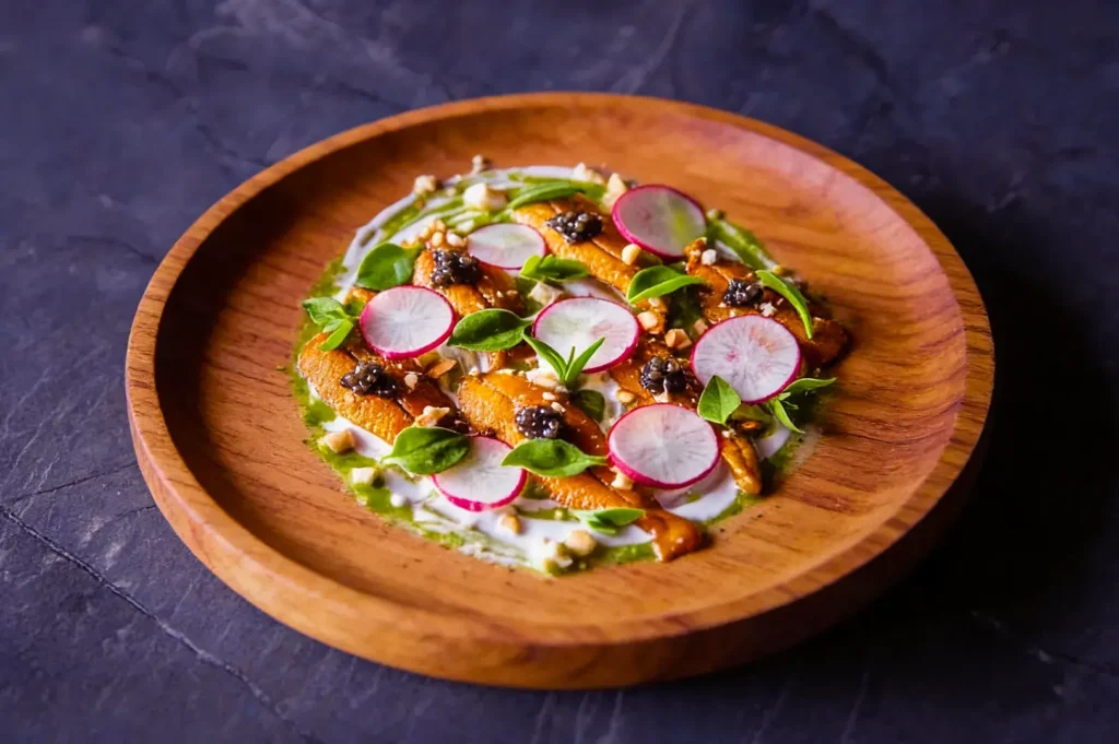 Wooden plate with a gourmet dish featuring sliced radishes, fresh greens, and dark garnishes on a creamy sauce, set against a dark background.
