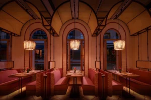Cozy restaurant interior with arched windows, soft pink booths, and elegant table settings. Warm pendant lights create an intimate, inviting ambiance.