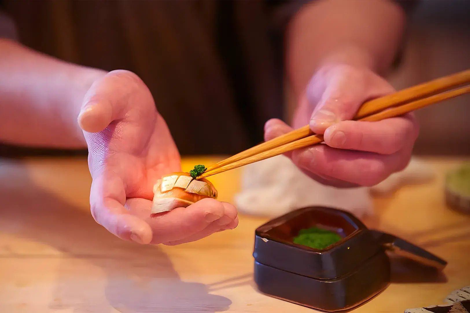 A person uses chopsticks to place sushi into their palm. The sushi appears artfully garnished. A small dish of green condiment is seen on the wooden table.