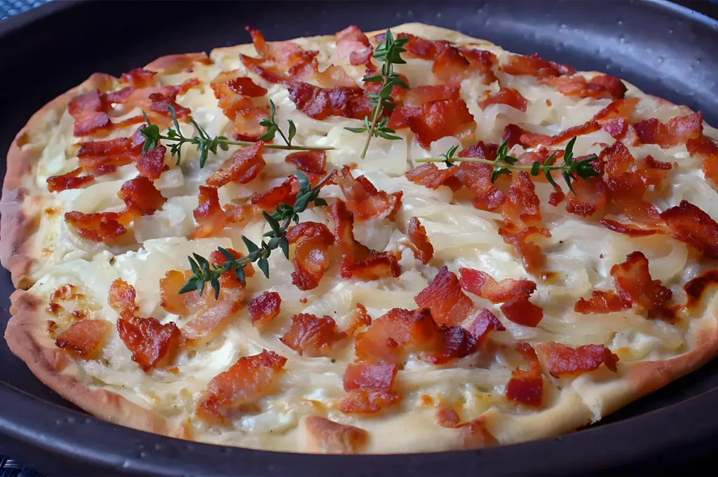 A savory tarte flambée on a dark tray, topped with crispy bacon, caramelized onions, and sprigs of fresh thyme, evoking a warm, rustic feel.