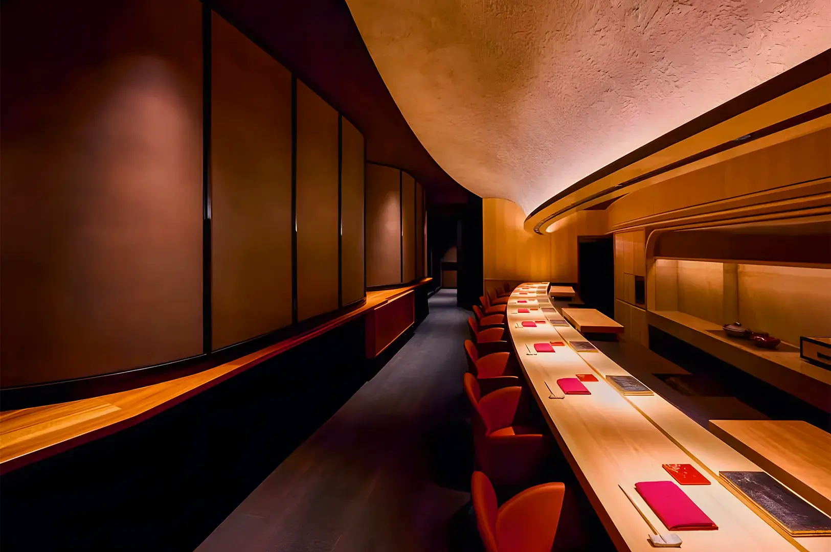 Elegant, dimly-lit restaurant interior with a long, curved wooden counter lined with red chairs and neatly placed red napkins, exuding a cozy ambiance.