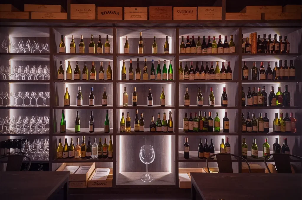 Dimly lit wine cellar with shelves full of assorted wine bottles and a large wine glass centered. Adjacent are rows of empty glasses, creating an elegant ambiance.