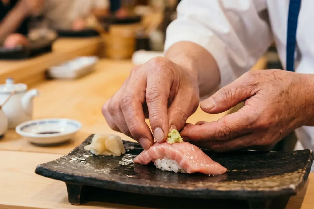Japanese chef hands placing wasabi on a piece of tuna nigiri sushi served on a black ceramic plate.