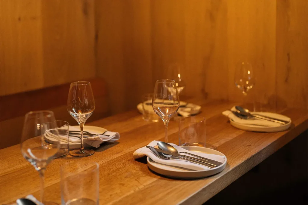 A neatly set wooden restaurant table with wine glasses, plates, and polished silverware in a warm, dimly lit dining room.