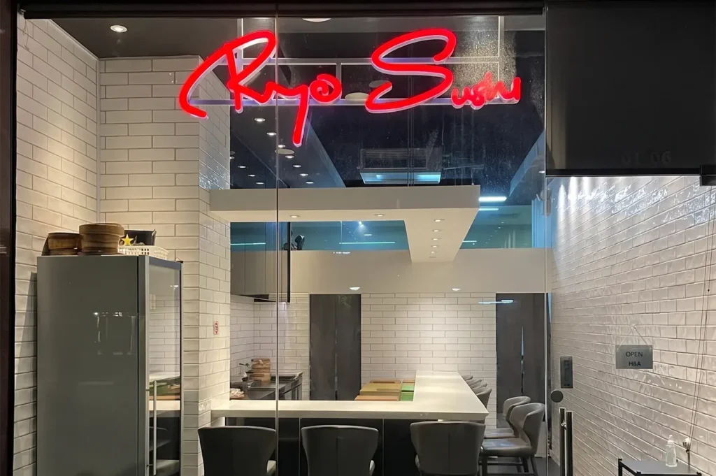 Modern glass storefront of Ryo Sushi, showcasing a vibrant red neon sign above an intimate dining area with a white L-shaped counter, grey bar stools, and white brick-patterned walls.