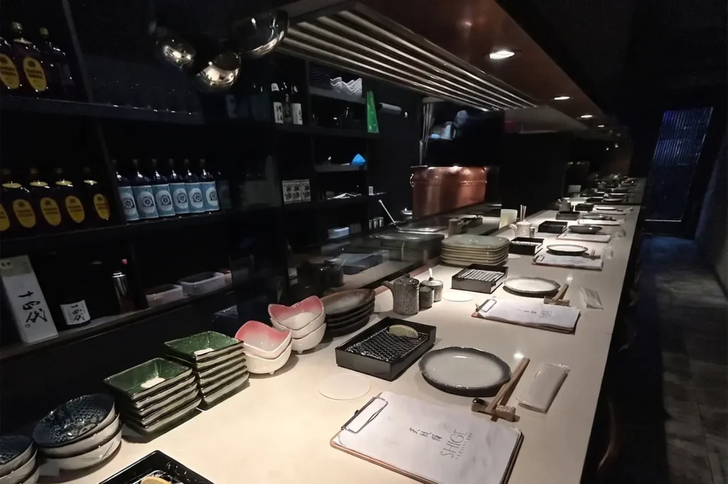 Tempura Shige restaurant long counter dining setup at Cuppage Plaza with organized tableware and sake bottles on display shelves.