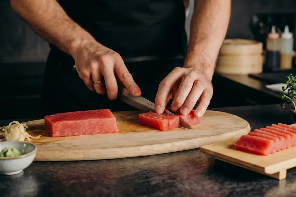 Chef slicing fresh raw tuna block into sashimi on a wooden cutting board.