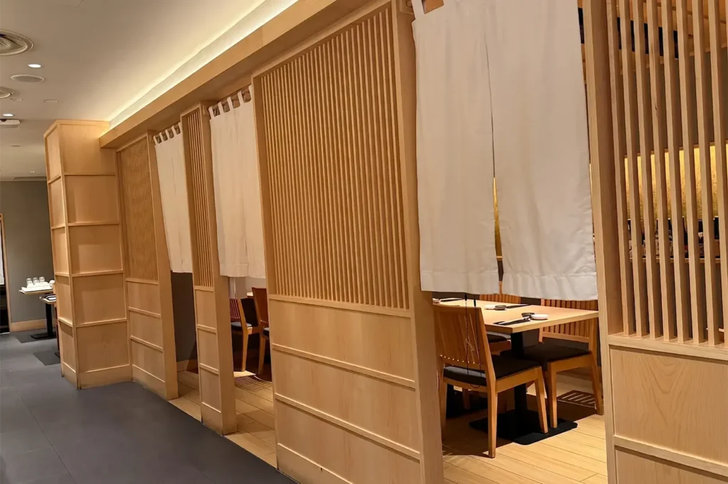 Traditional wooden booth seating at Haku Sushi, featuring light-wood lattice partitions, white noren curtains, and minimalist dining tables for private dining.