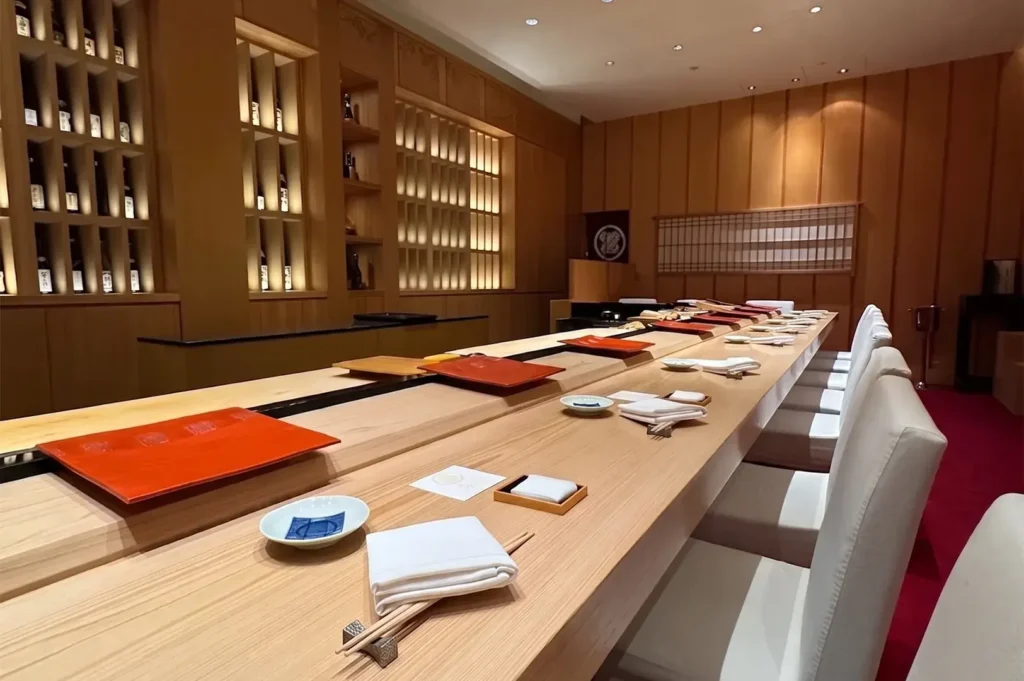 Elegant dining interior at Shinji by Kanesaka, featuring a minimalist wooden sushi counter with neatly arranged place settings and light-wood paneling.