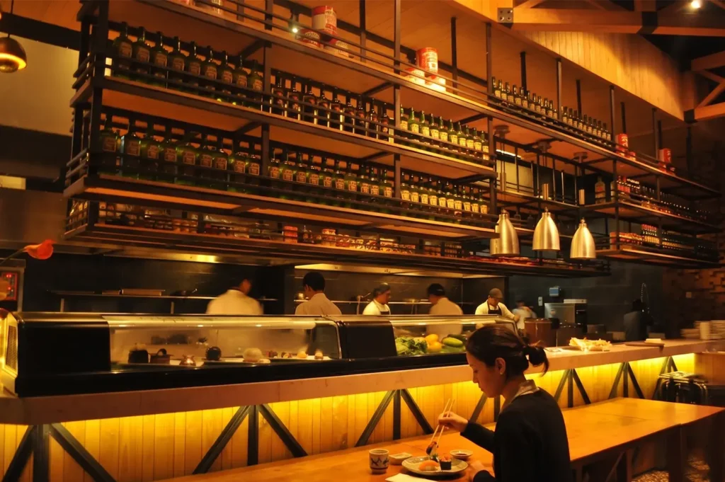 A woman eating sushi at a modern restaurant counter with chefs working in an open kitchen background.