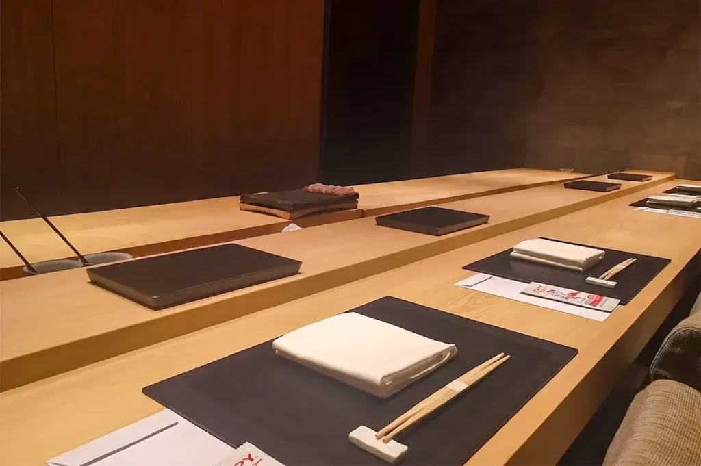 Minimalist wooden sushi counter at Shoukouwa, showing immaculate place settings with chopsticks, white napkins on black placemats, and a dim, intimate dining ambiance.