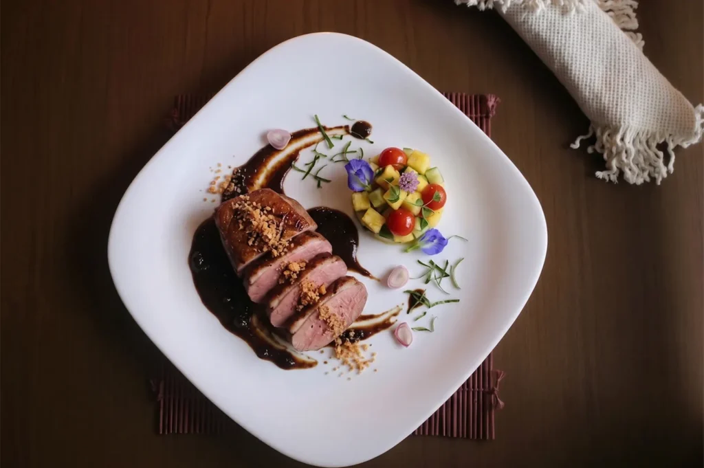Sliced roasted duck breast with dark sauce and mango tomato salsa on a white plate.