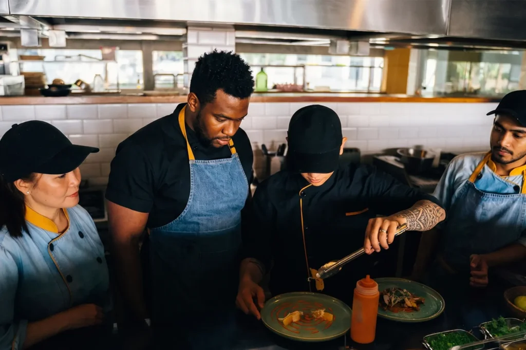 A diverse team of chefs in uniforms watching a cook carefully plate a dish in a professional kitchen.