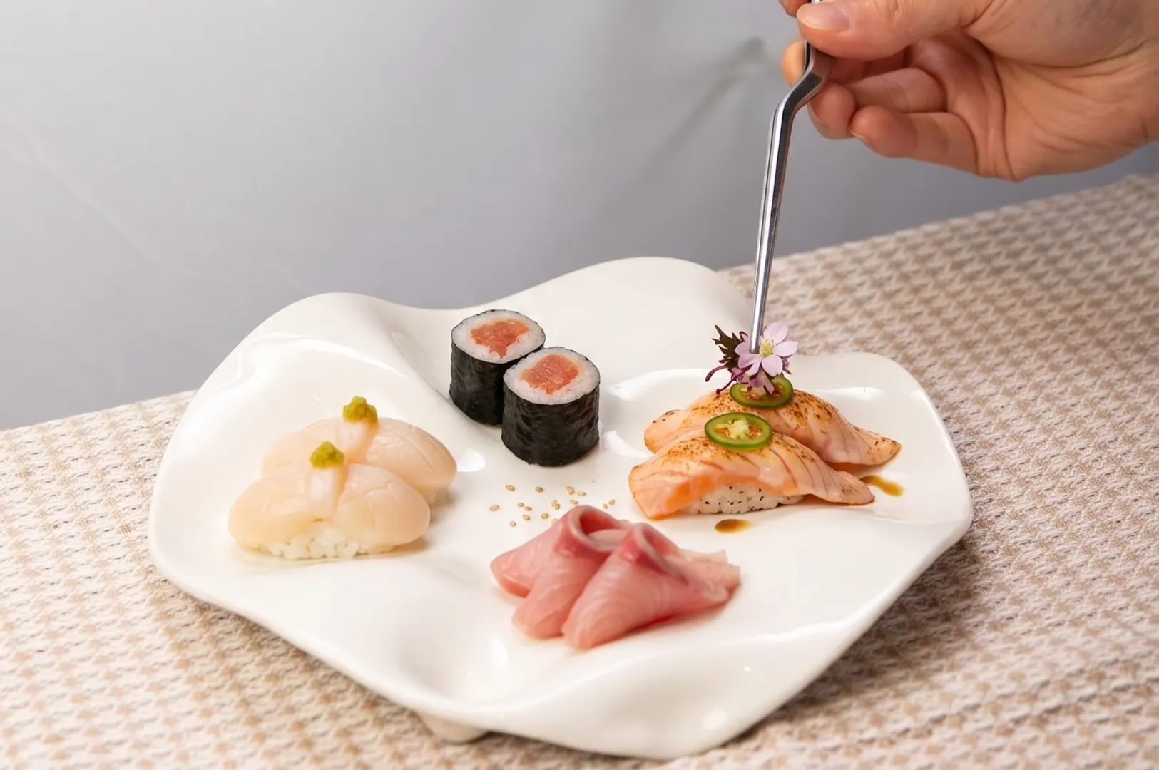 A hand uses tweezers to garnish a plate of assorted sushi, including nigiri, maki, and sashimi, served on a modern white platter.