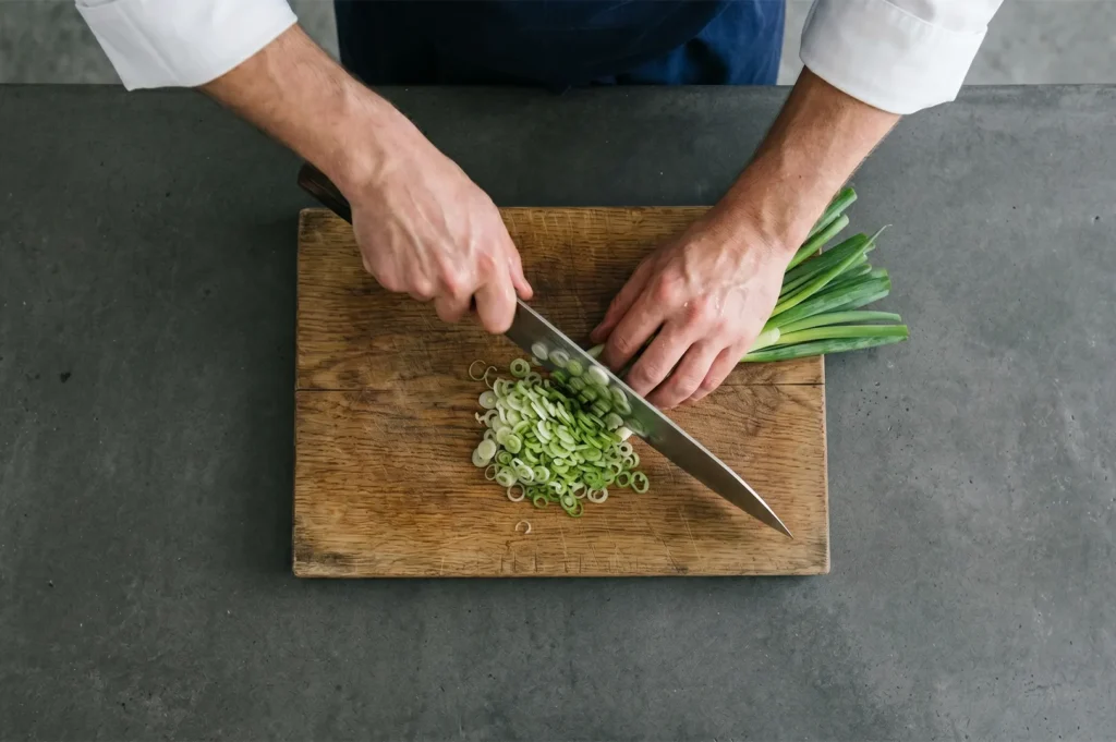 A professional chef in a white uniform chops fresh green onions on a wooden cutting board.