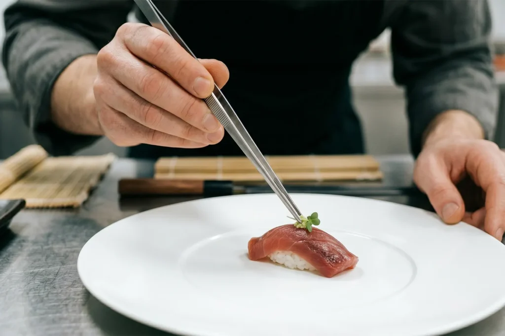 Chef using tweezers to garnish a piece of tuna nigiri sushi on a white plate.
