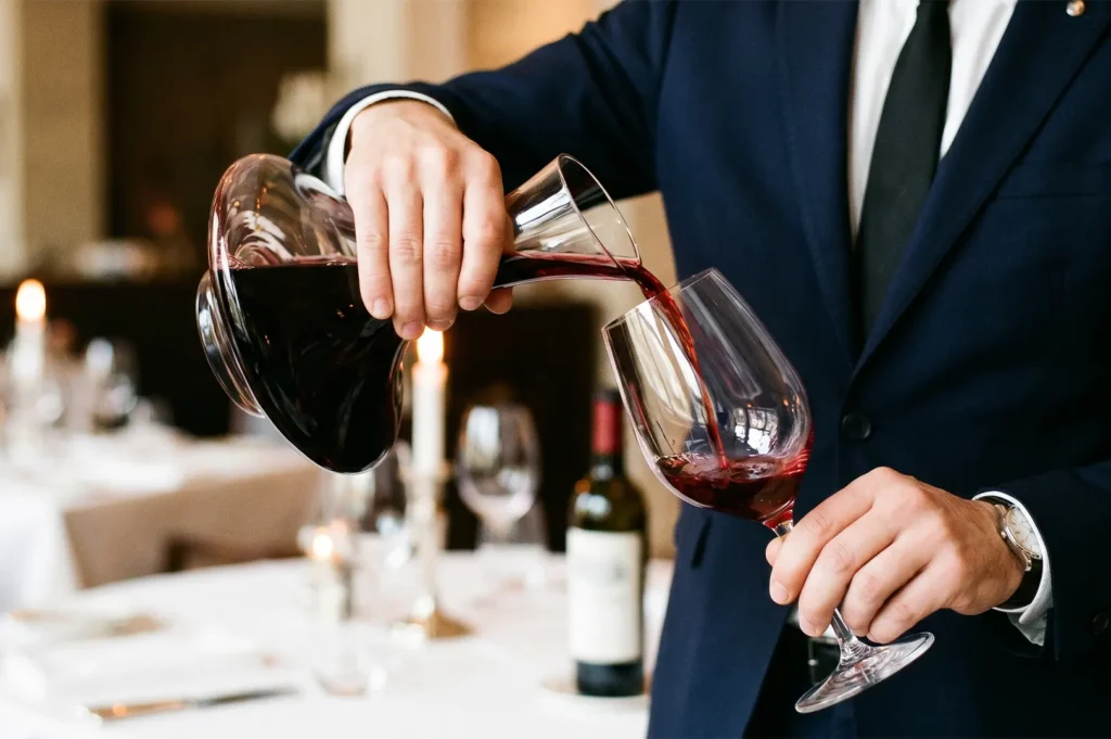 Sommelier in a suit pouring red wine from a glass decanter into a wine glass.