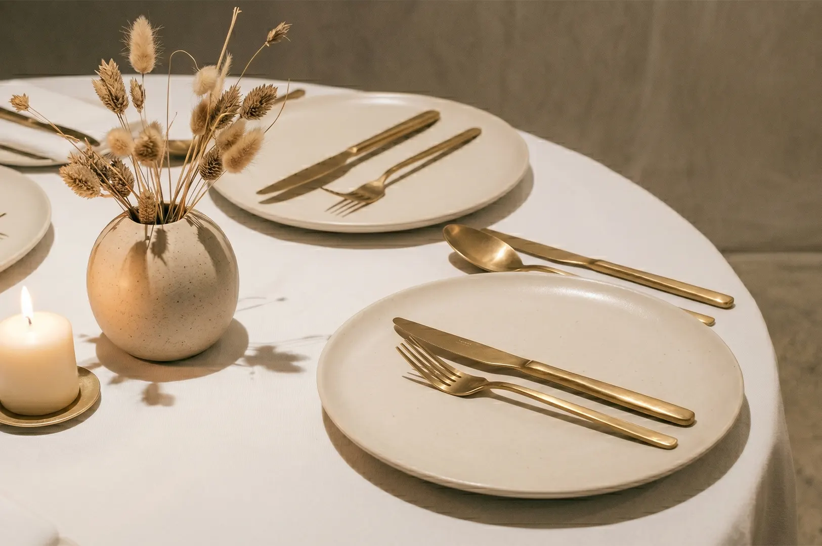 Minimalist table setting with matte ceramic plates, gold cutlery, and a small vase of dried rabbit tail grass.