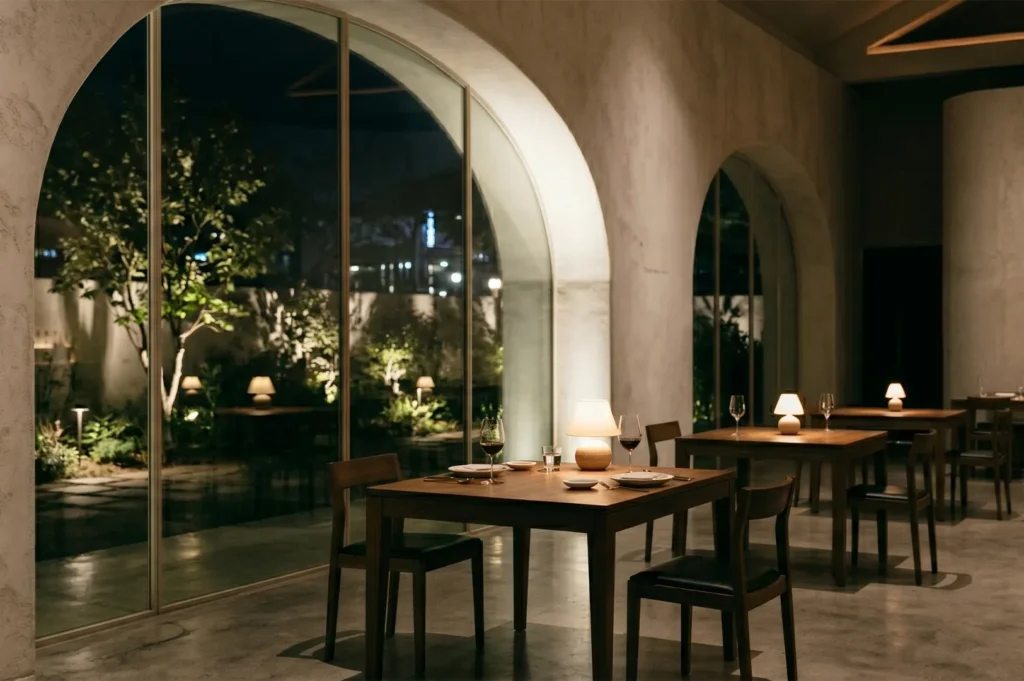 Minimalist fine dining restaurant interior featuring wooden tables, warm lamps, and large arched windows looking into a garden at night.