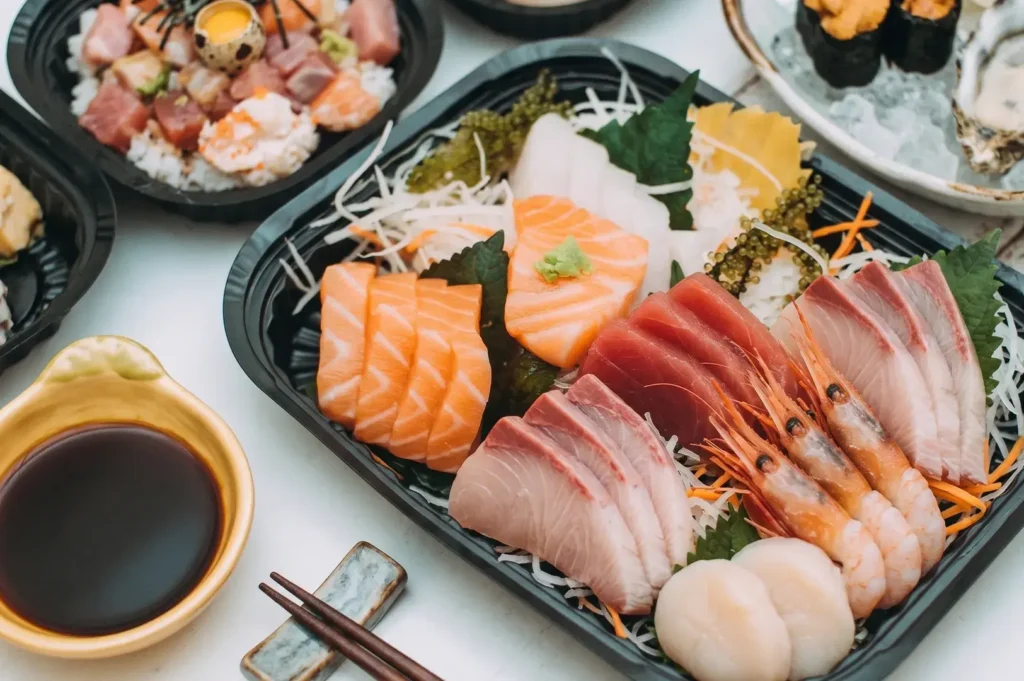 Fresh sashimi platter with salmon, tuna, hamachi yellowtail, sweet shrimp, and scallops in a takeout container.