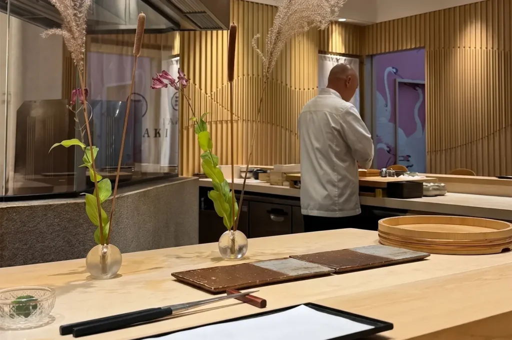 Chef preparing sushi at the elegant counter of Sushi Masaaki, featuring minimalist wooden decor, delicate ikebana floral arrangements, and a serene, upscale dining atmosphere.
