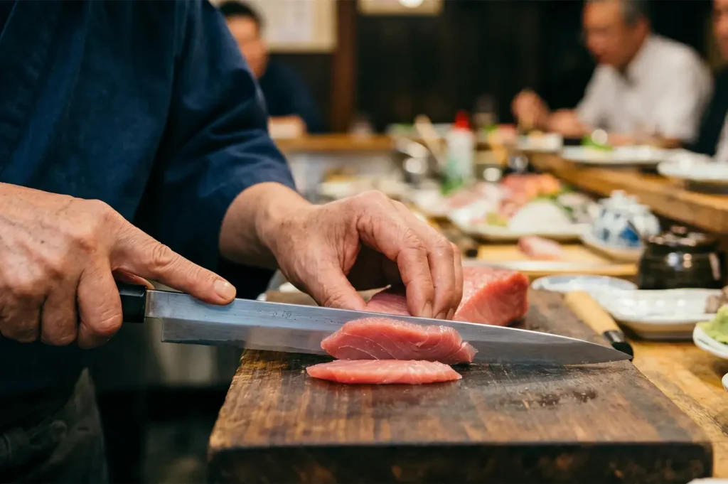 Japanese sushi chef slicing fresh raw tuna sashimi with a traditional yanagiba knife on a wooden cutting board.