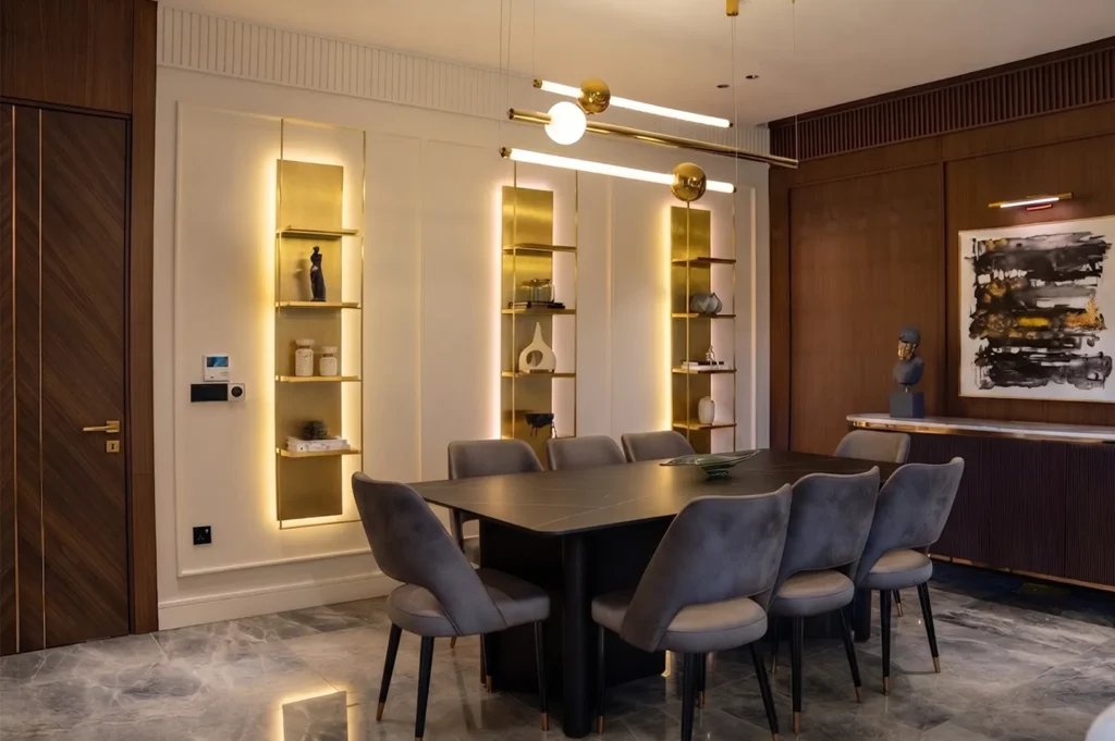 Modern luxury dining room featuring a black marble table, grey velvet chairs, and illuminated gold wall shelving.