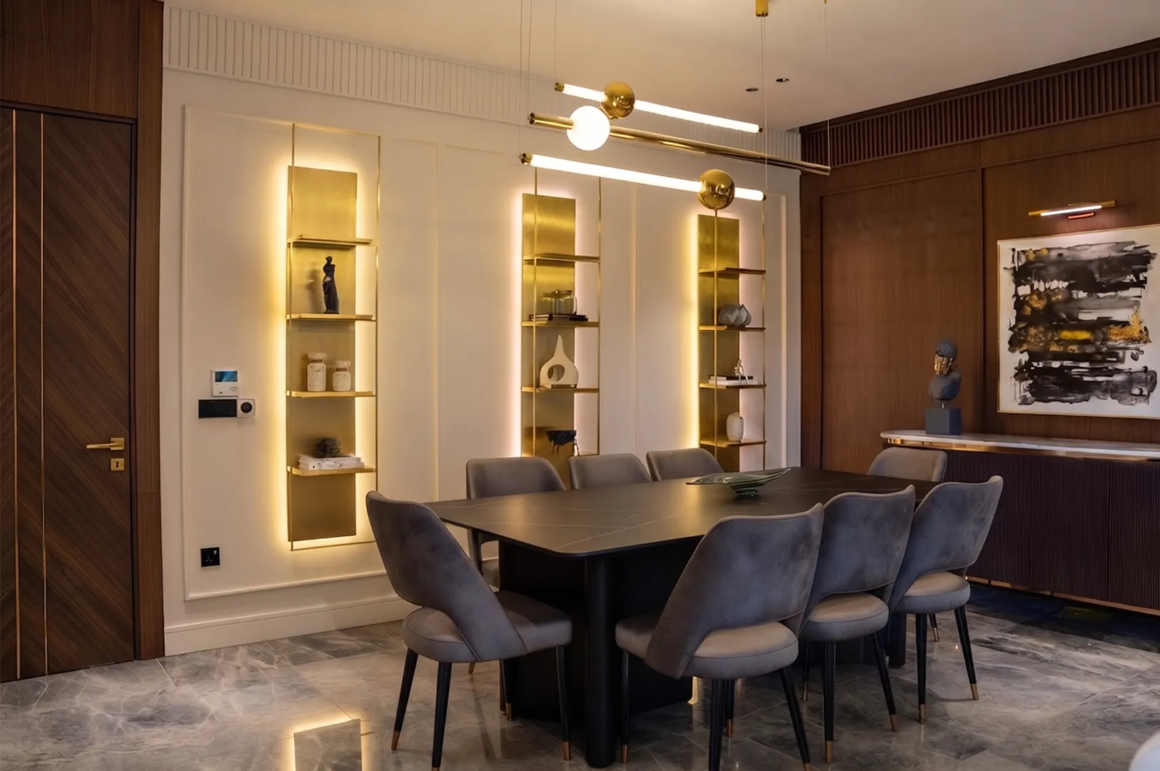 Modern luxury dining room featuring a black marble table, grey velvet chairs, and illuminated gold wall shelving.